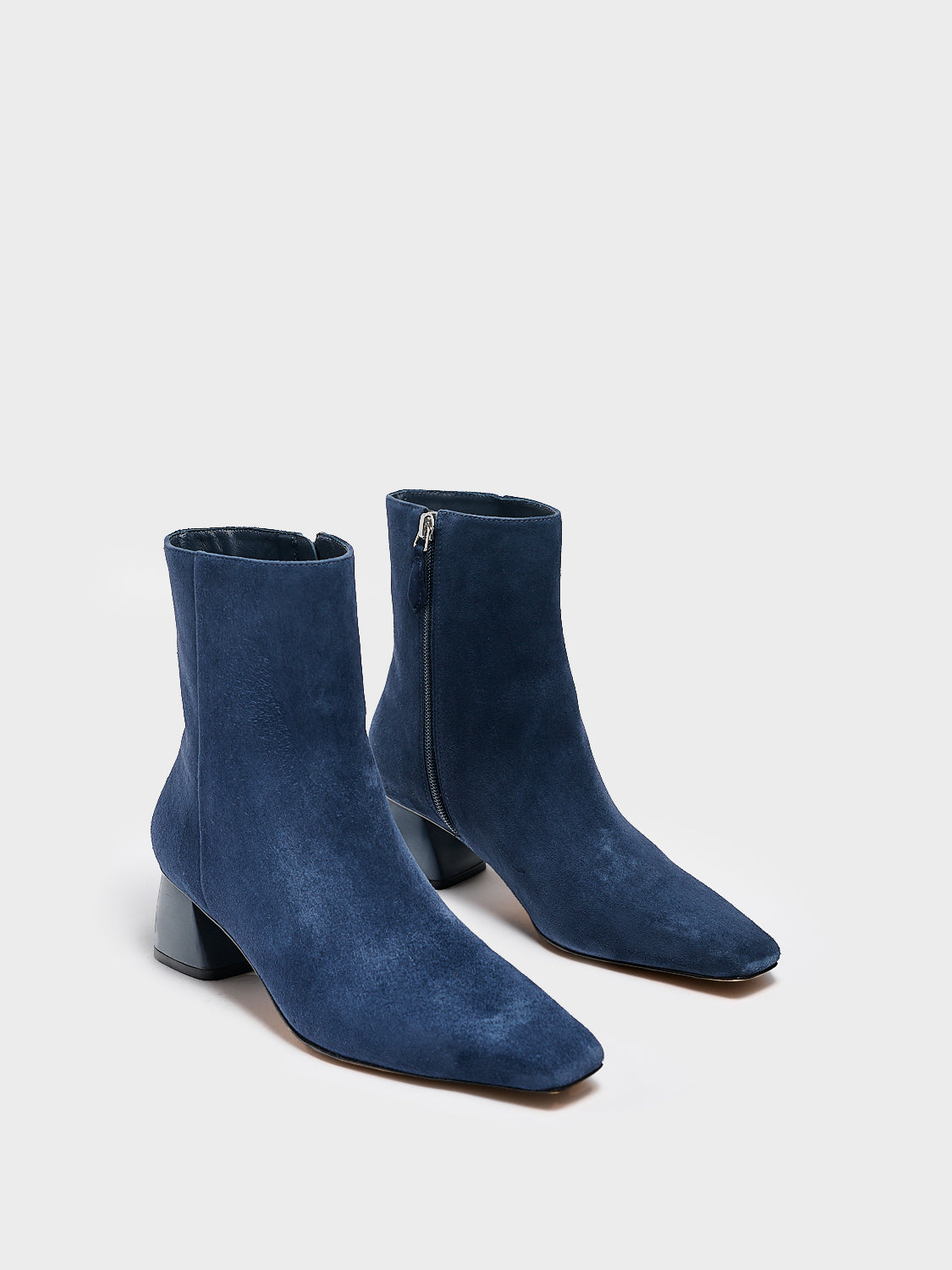 Angle view of a pair of navy suede ankle booties with a rounded heel shape, zipper closure, and square toe.