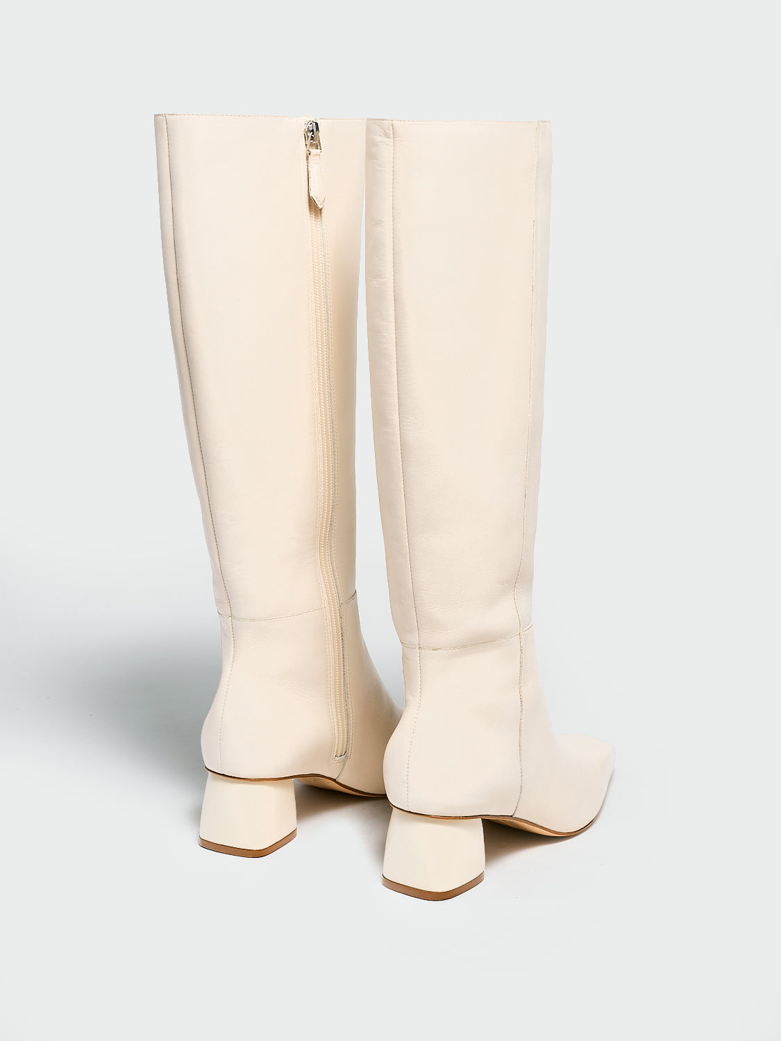 Back view of a pair of ivory nappa leather below the knee tall boots with a rounded heel shape, zipper closure, and square toe.