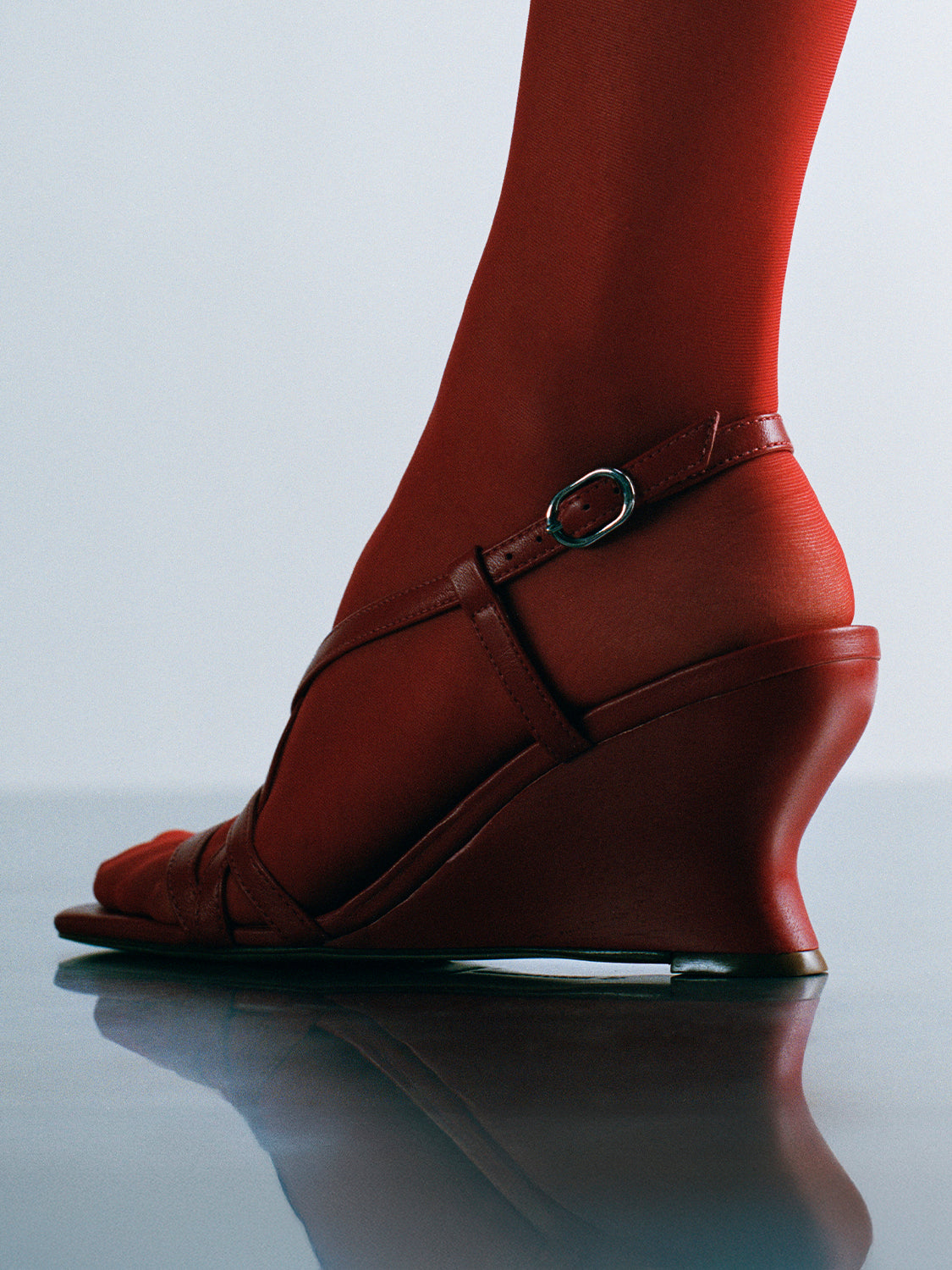 Profile view of a lipstick red leather strappy sandal with a sculptural wedge, shown on a model.