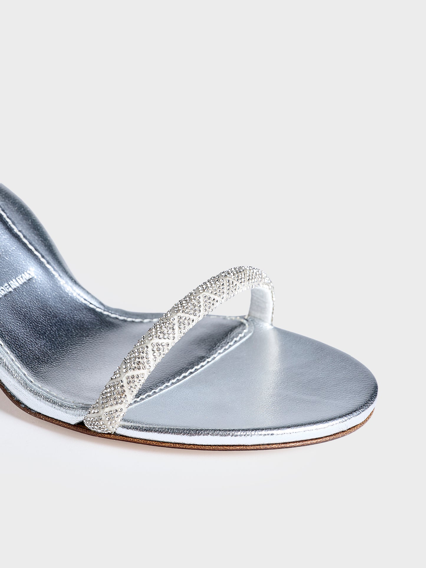 Detail view of a pair of silver metallic leather high heel sandals with a crystal embellished toe strap and adjustable ankle strap.