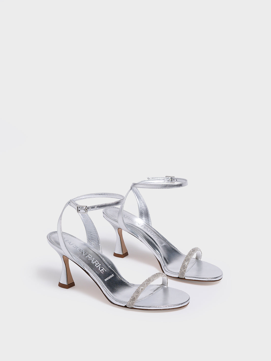 Angle view of a pair of silver metallic leather high heel sandals with a crystal embellished toe strap and adjustable ankle strap.