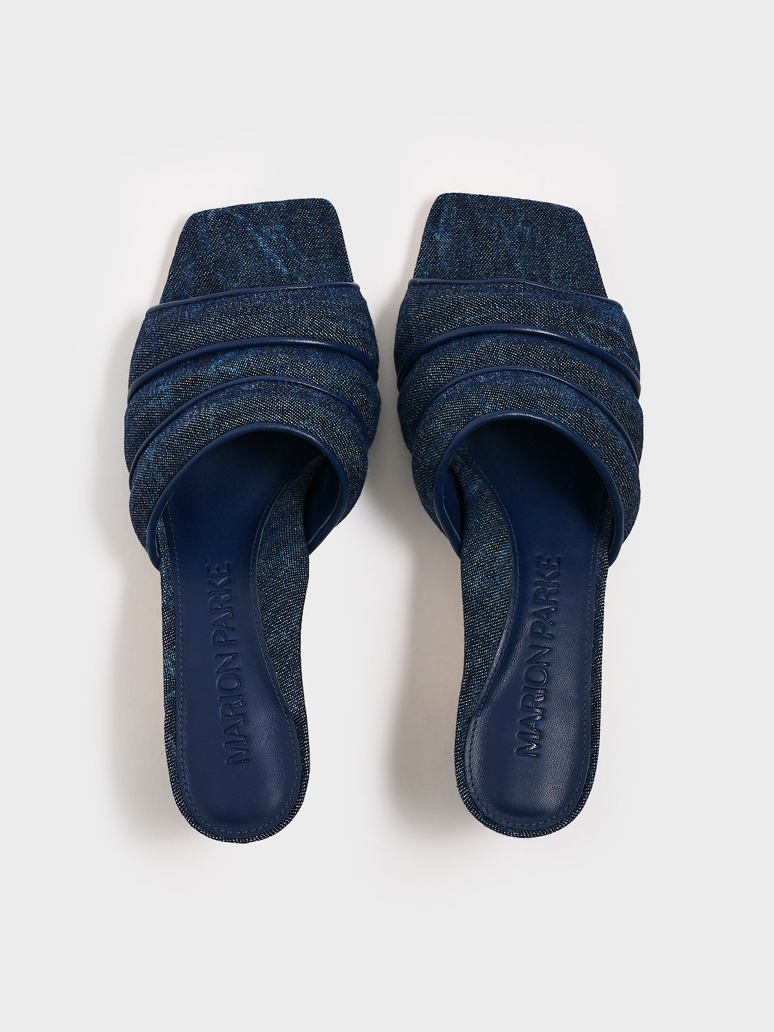 Top view of a pair of blue denim slide kitten heels with a leather trimed puffy strap detail.