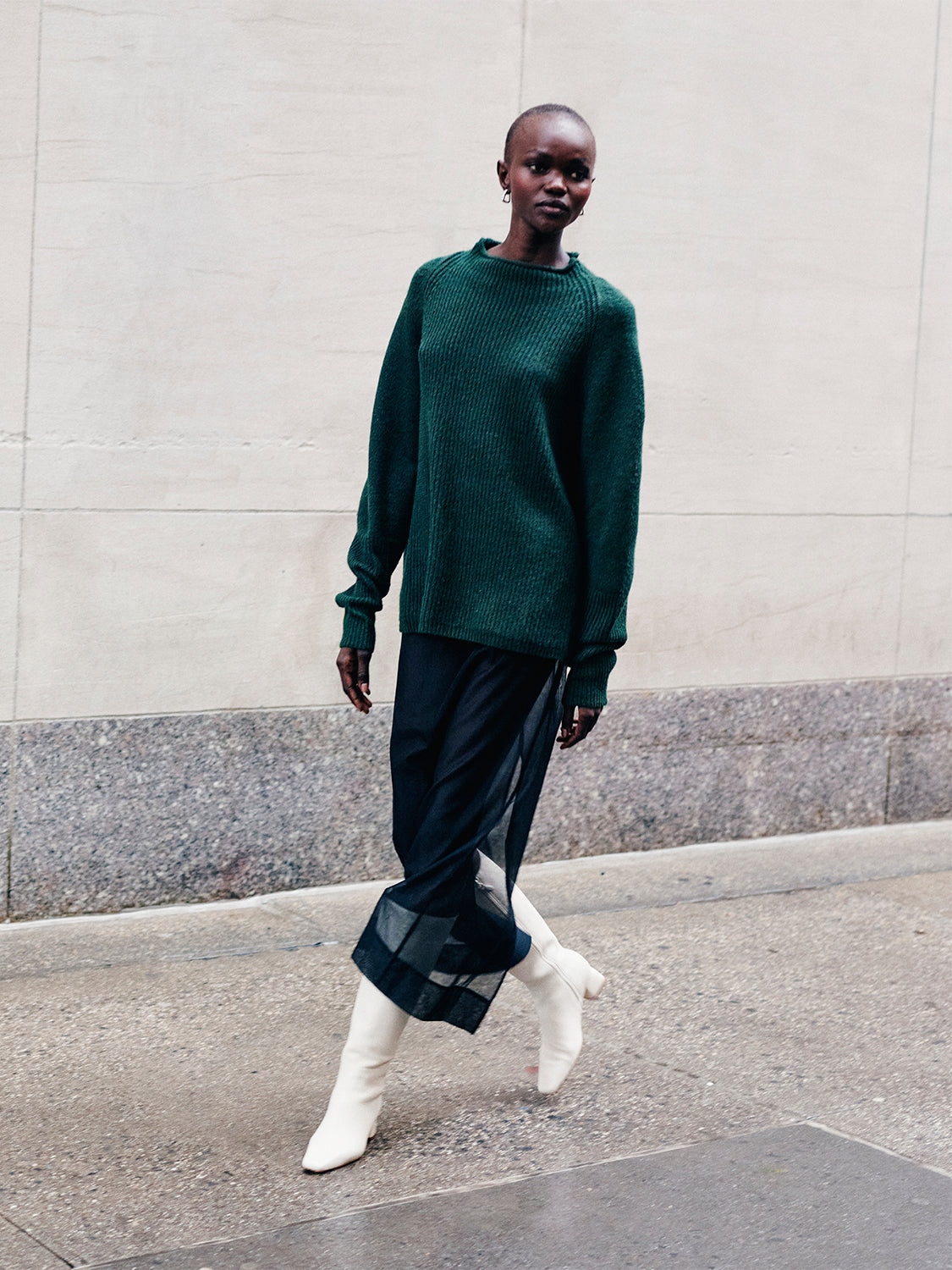 Woman wearing a green sweater and black sheer skirt with ivory tall boots walking down the street. 