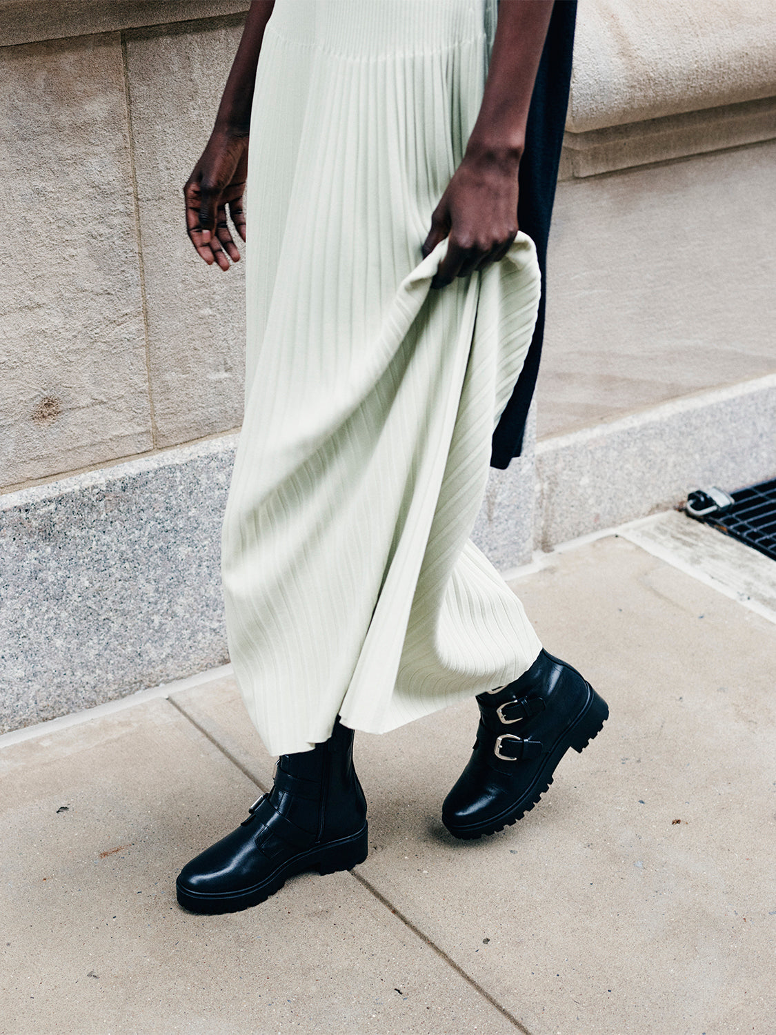 Model wearing a green dress, black scarf pleated dress standing against a stone building.