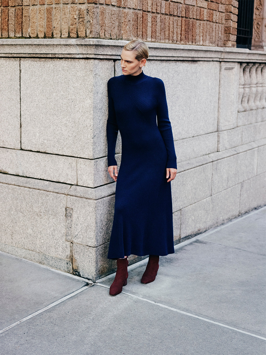 Woman in a navy blue dress and merlot suede ankle boots, standing against a stone wall.