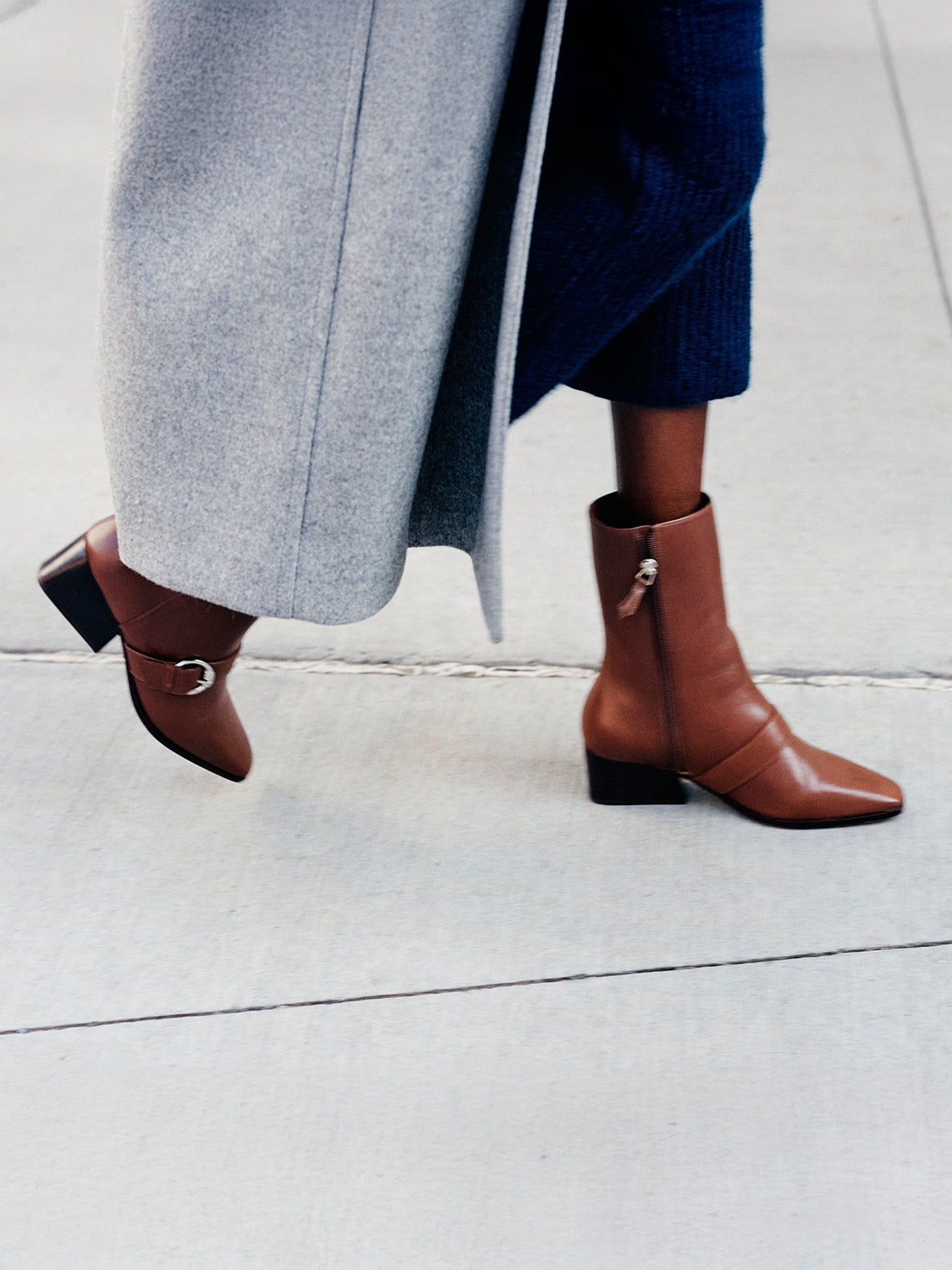 Woman wearing a blue sweater skirt set with a grey wool coat and brown boots, walking on the sidewalk.