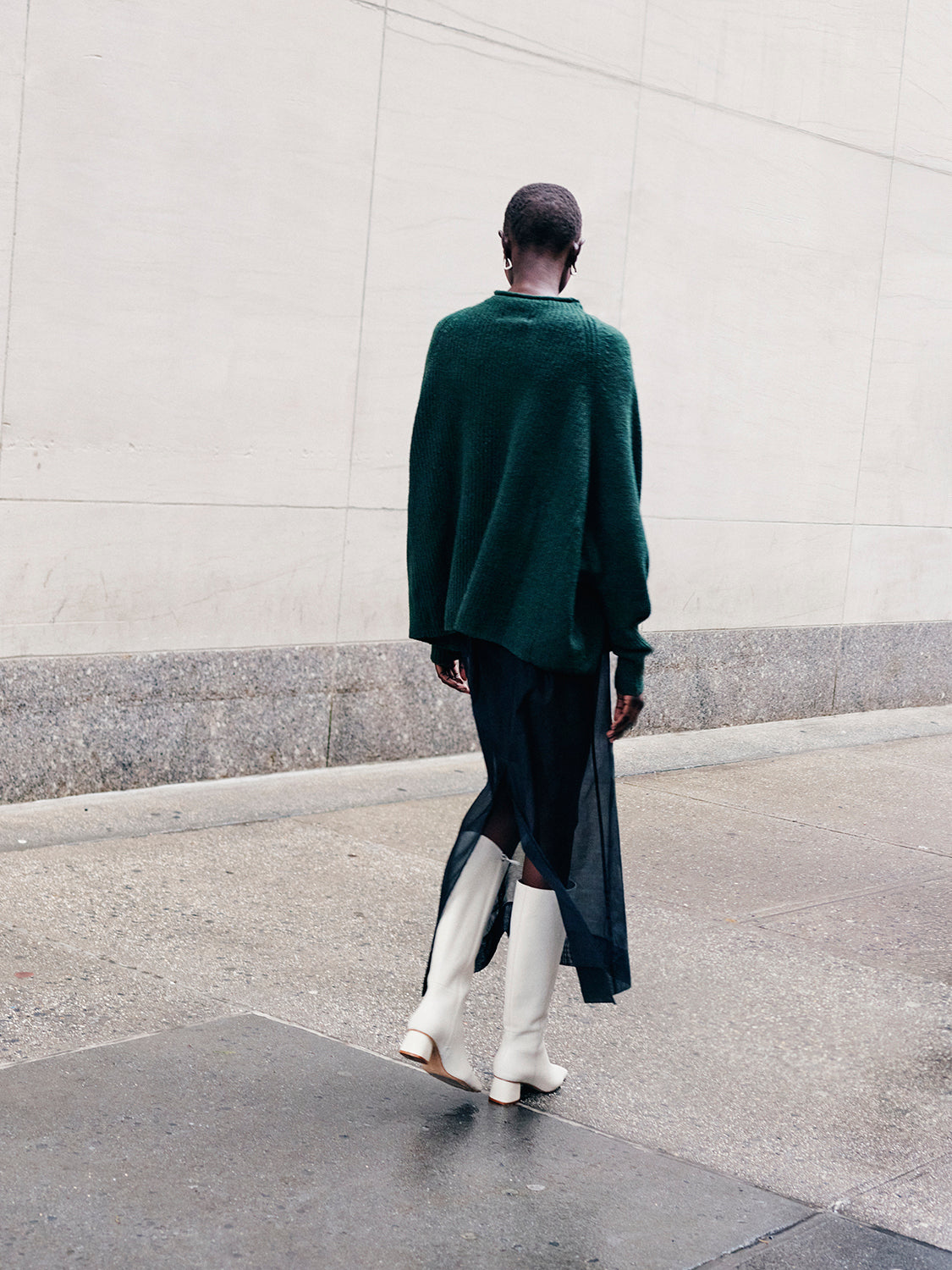 Woman wearing a green sweater and black sheer skirt with ivory tall boots walking down the street. 