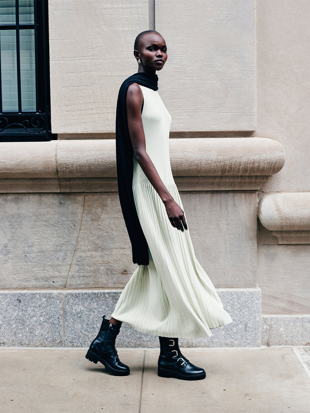 Model wearing a green dress, black scarf pleated dress standing against a stone building.
