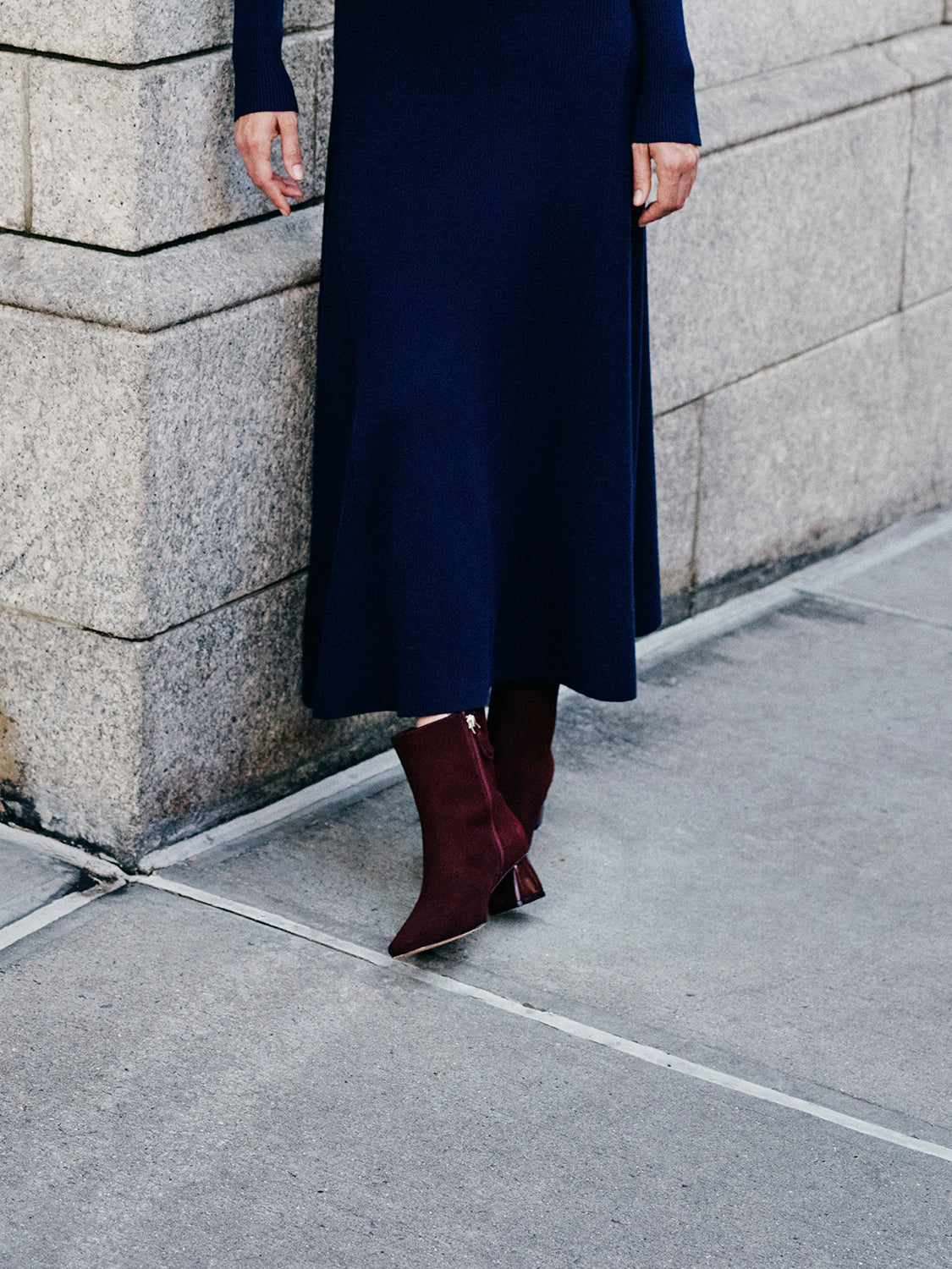 Woman wearing a navy dress and merlot suede ankle boots, standing on a street corner. 