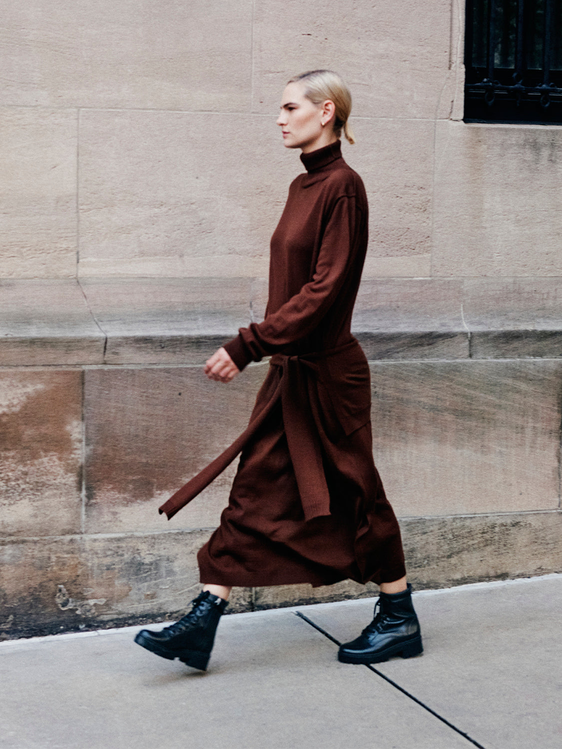 Woman wearing a brown dress and black lug sole lace-up boots walking on a sidewalk. 