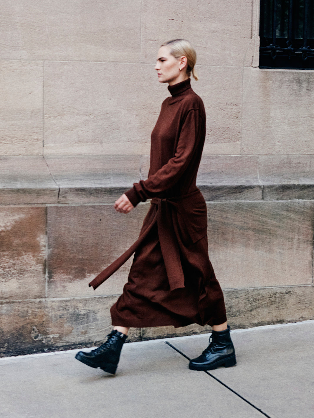 Woman wearing a brown dress and black lug sole lace-up boots walking on a sidewalk. 