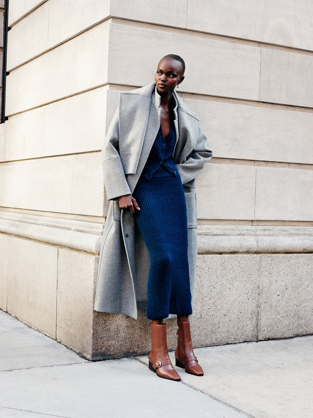 Woman wearing a blue sweater skirt set with a grey wool coat and brown boots, leaning against a wall.