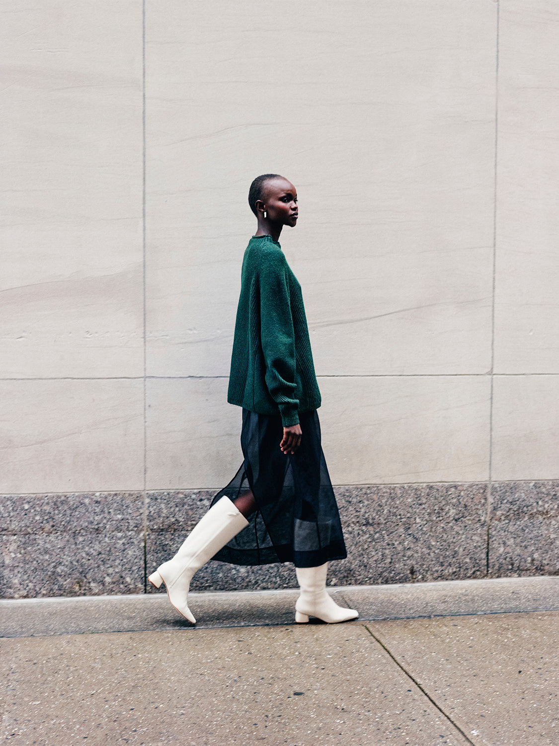 Woman wearing a green sweater and black sheer skirt with ivory tall boots walking down the street. 