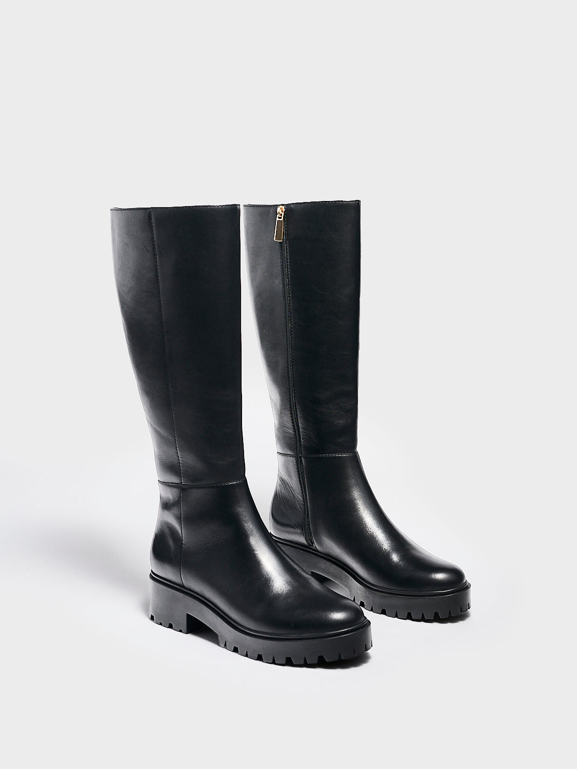 Angle view of a pair of black nappa leather tall boots with a lug sole, zipper closure, and round toe. 