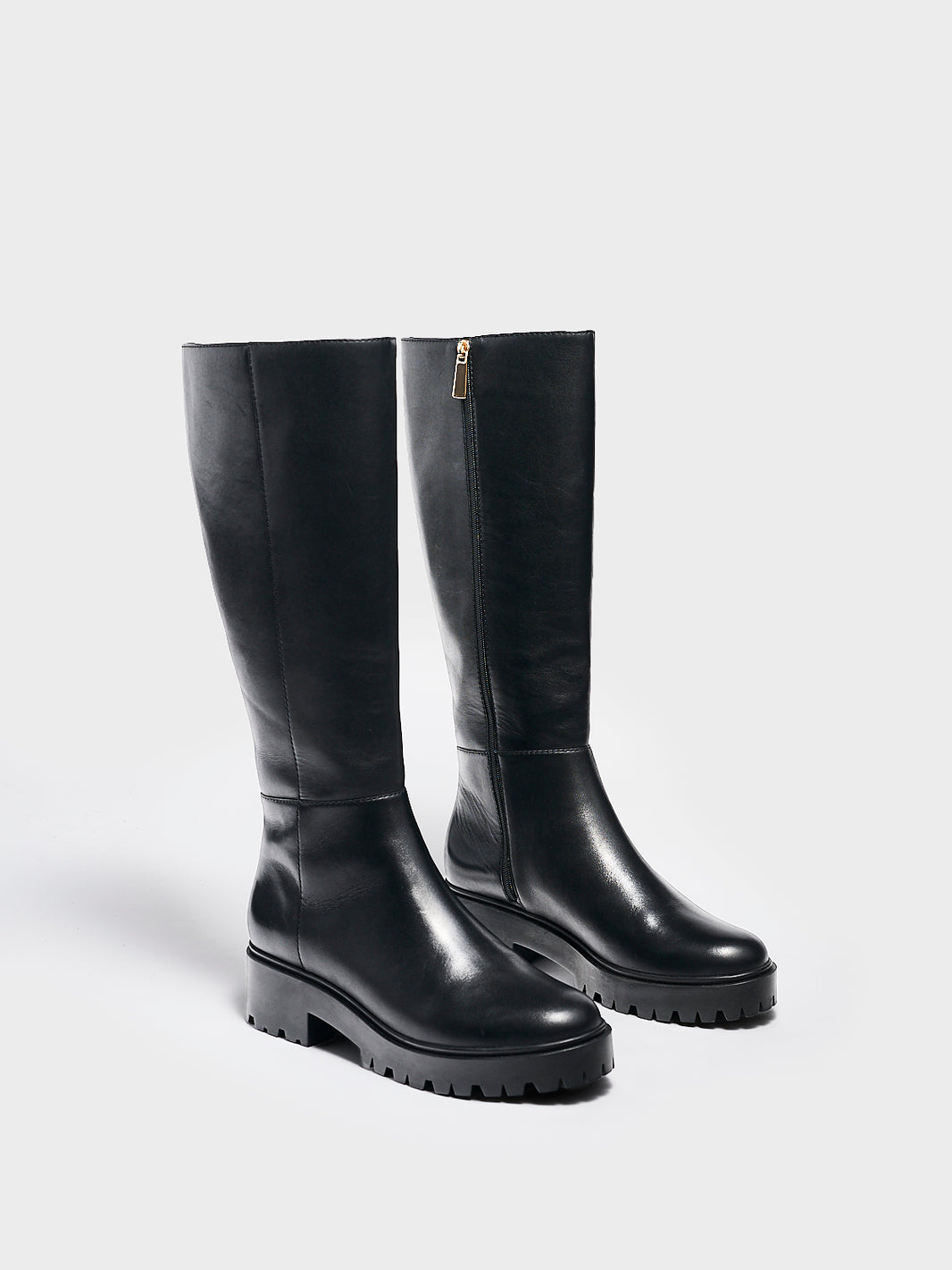 Angle view of a pair of black nappa leather tall boots with a lug sole, zipper closure, and round toe. 