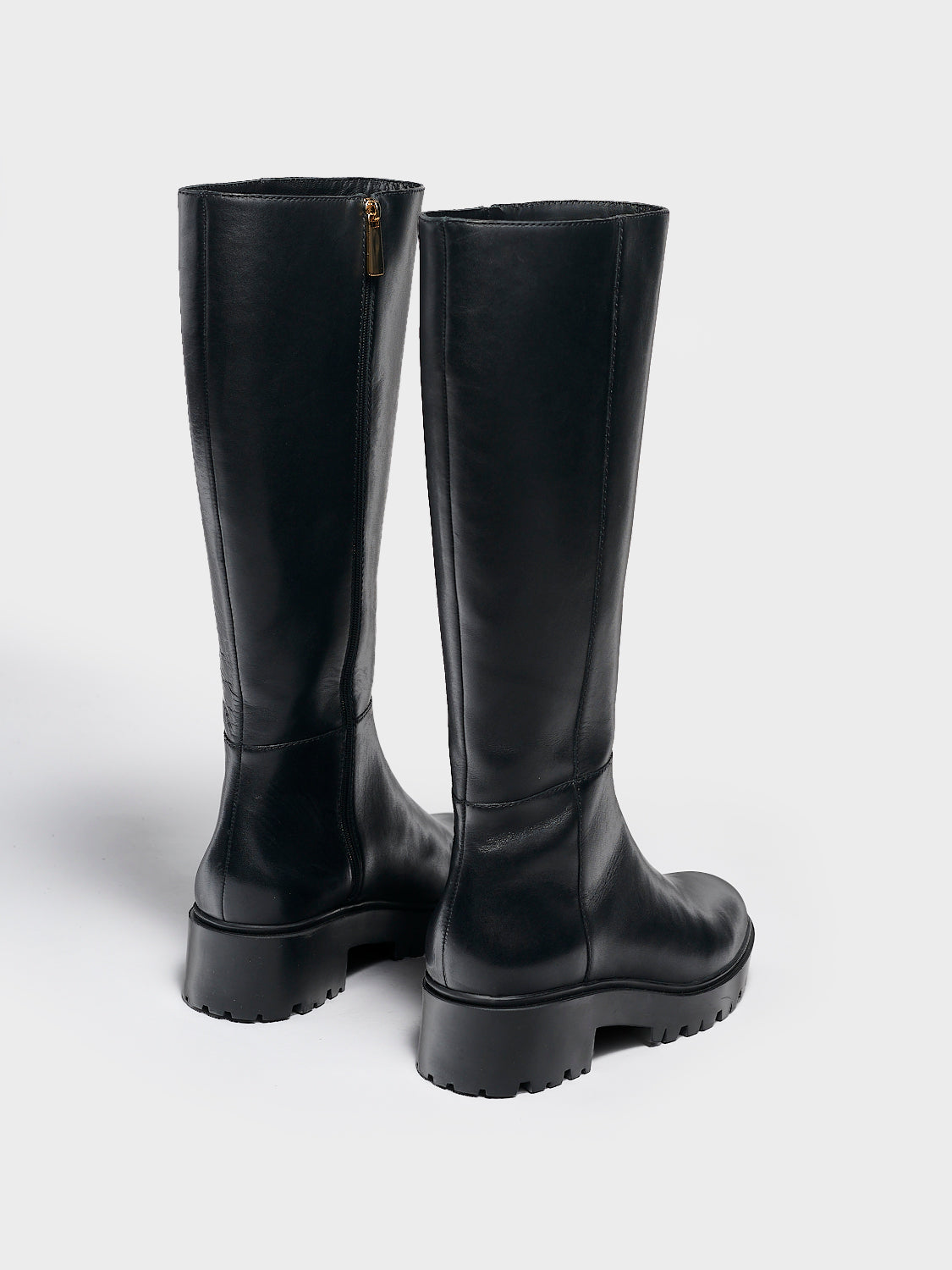 Back view of a pair of black nappa leather tall boots with a lug sole, zipper closure, and round toe. 
