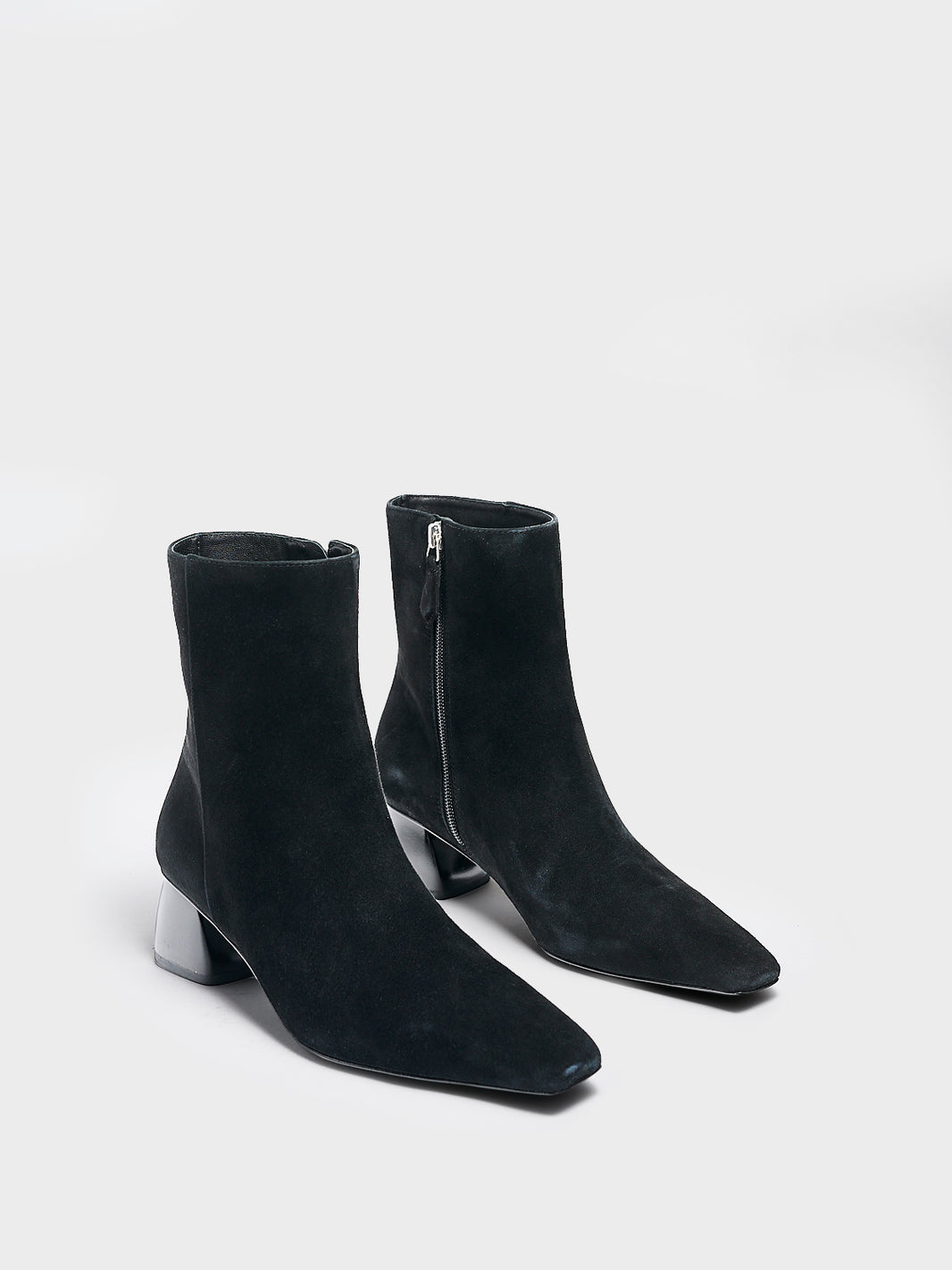 Angle view of a pair of black suede ankle booties with a rounded heel shape, zipper closure, and square toe. 