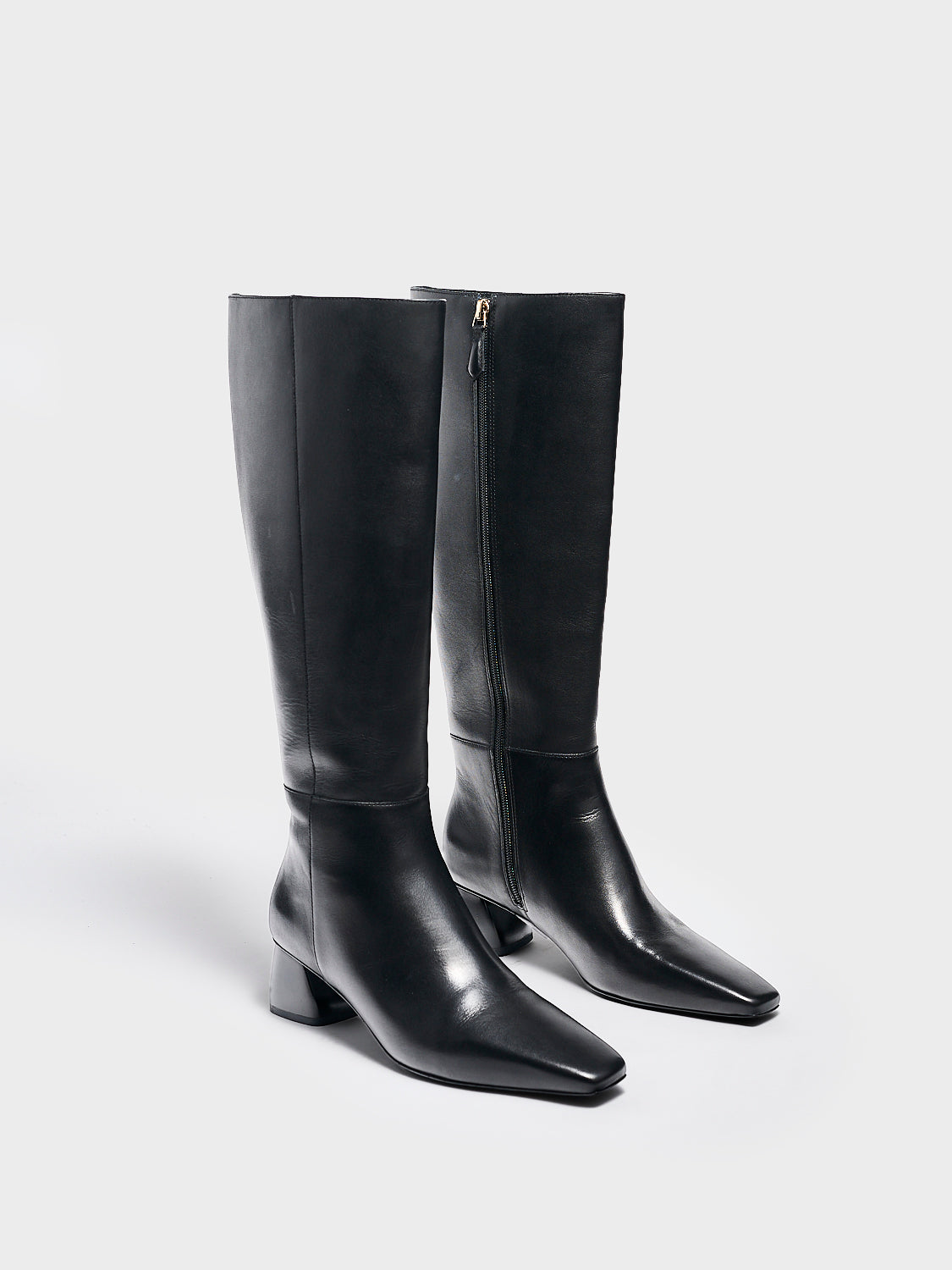 Angle view of a pair of black nappa leather below the knee tall boots with a rounded heel shape, zipper closure, and square toe. 