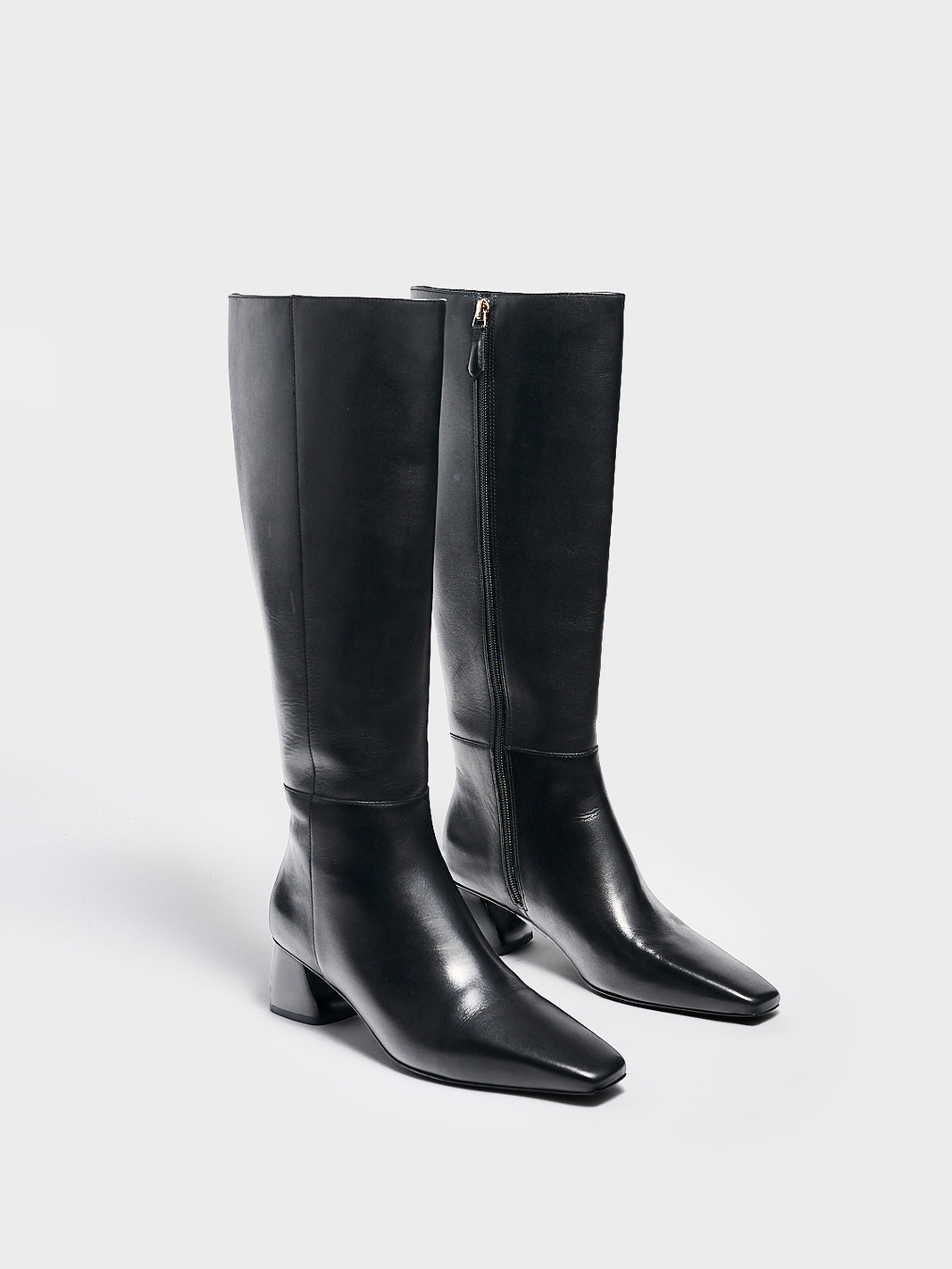 Angle view of a pair of black nappa leather below the knee tall boots with a rounded heel shape, zipper closure, and square toe. 