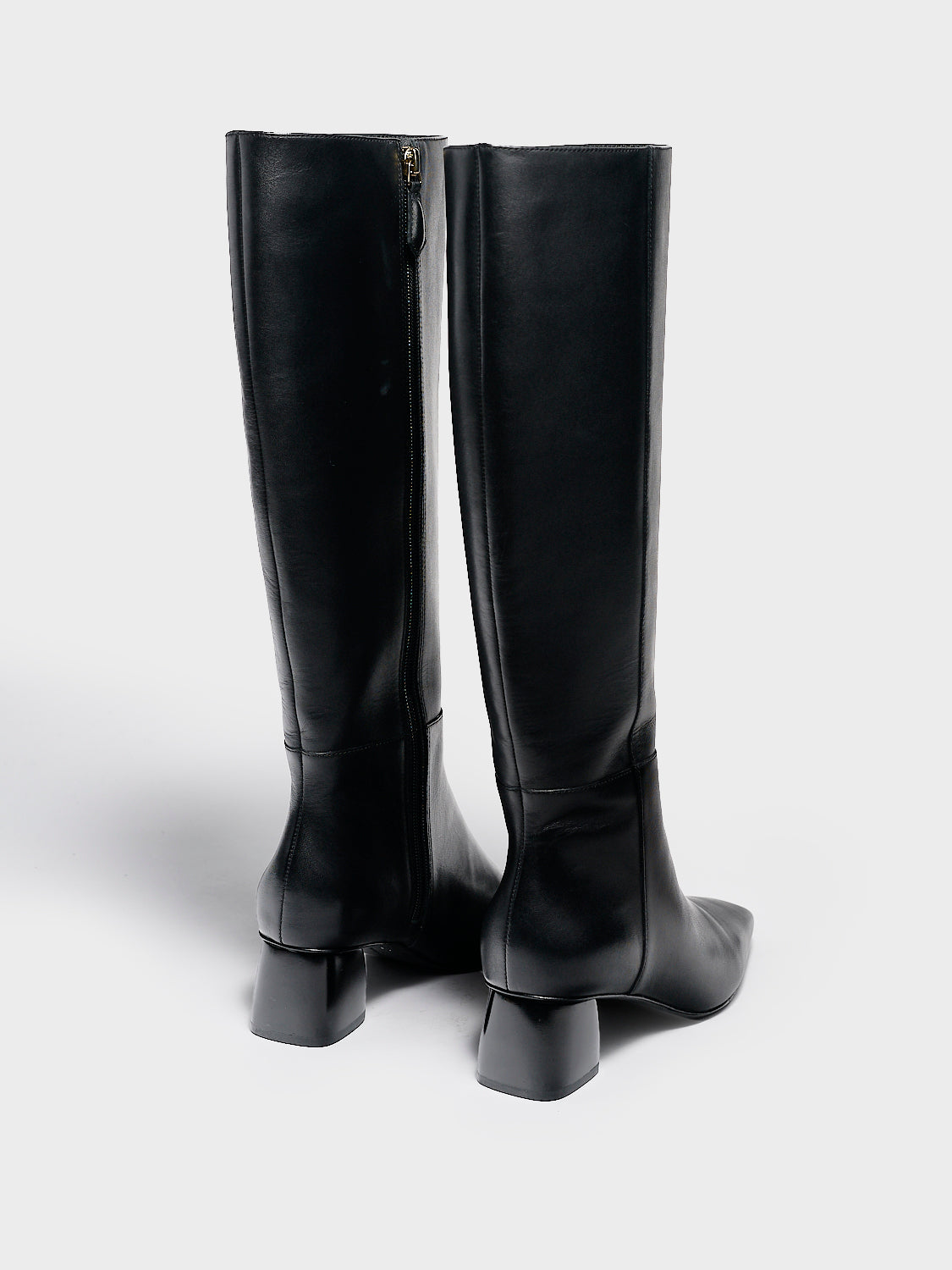Back view of a pair of black nappa leather below the knee tall boots with a rounded heel shape, zipper closure, and square toe. 