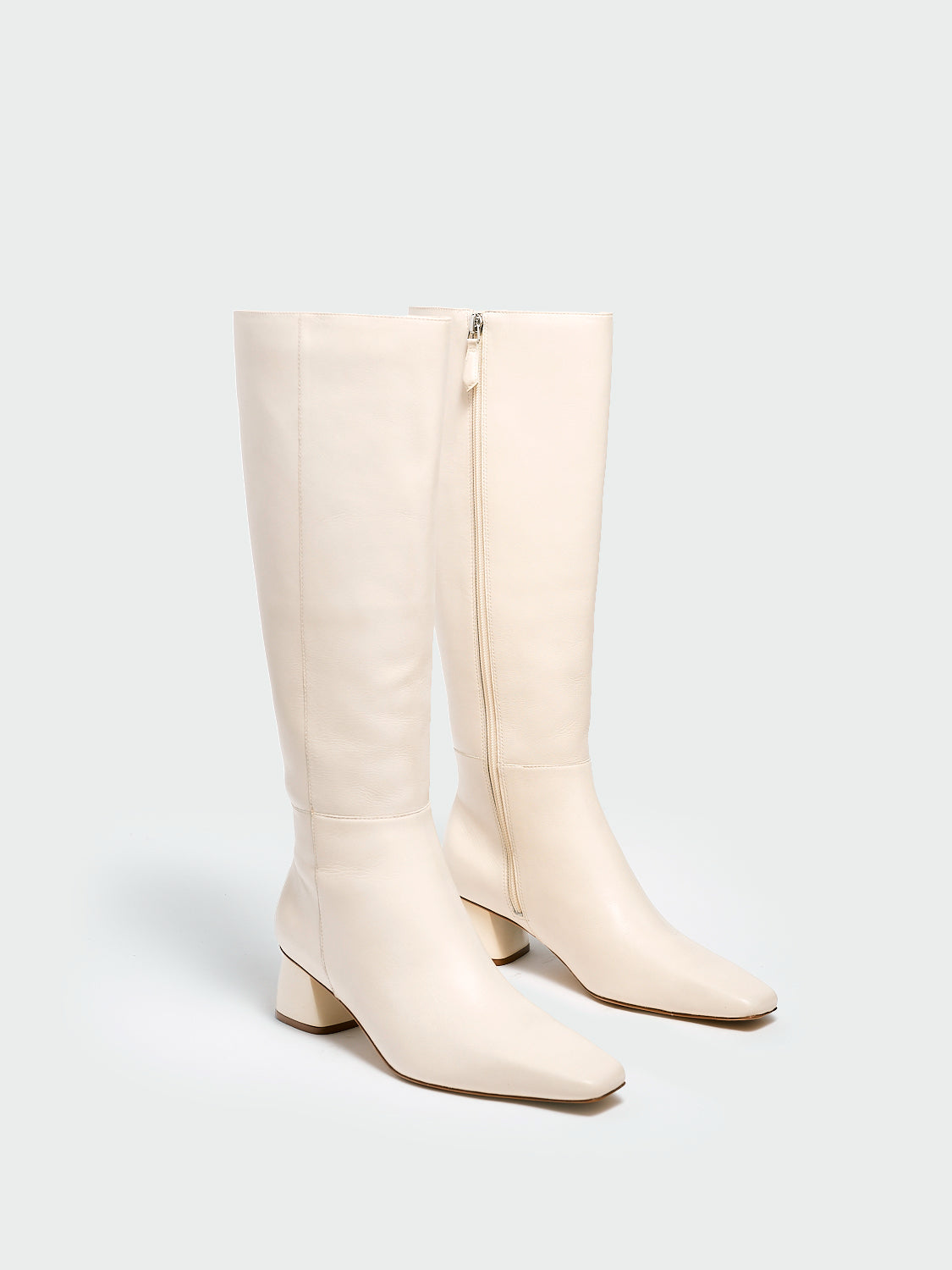 Angle view of a pair of ivory nappa leather below the knee tall boots with a rounded heel shape, zipper closure, and square toe. 