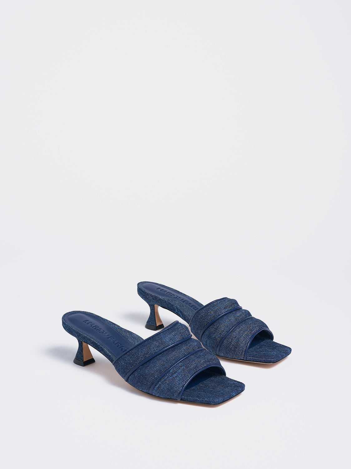 Angle view of a pair of blue denim slide kitten heels with a leather trimed puffy strap detail.
