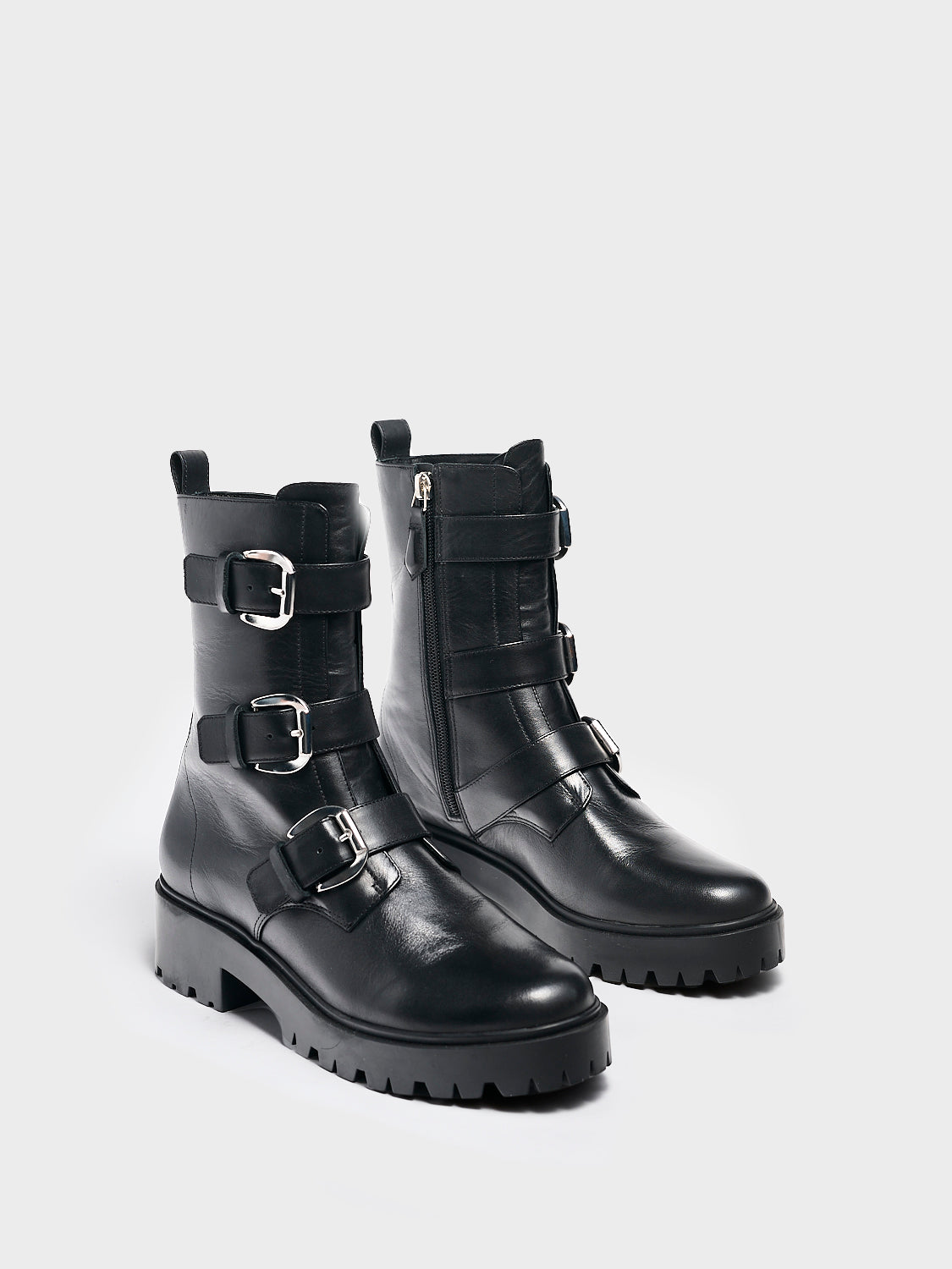Angle view of a pair of black nappa leather moto boots with a lug sole, zipper closure, silver buckles, and round toe. 