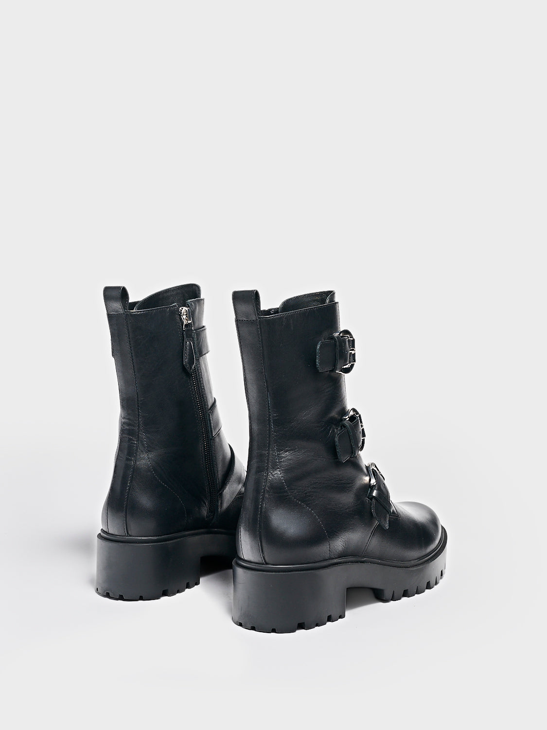 Back view of a pair of black nappa leather moto boots with a lug sole, zipper closure, silver buckles, and round toe. 