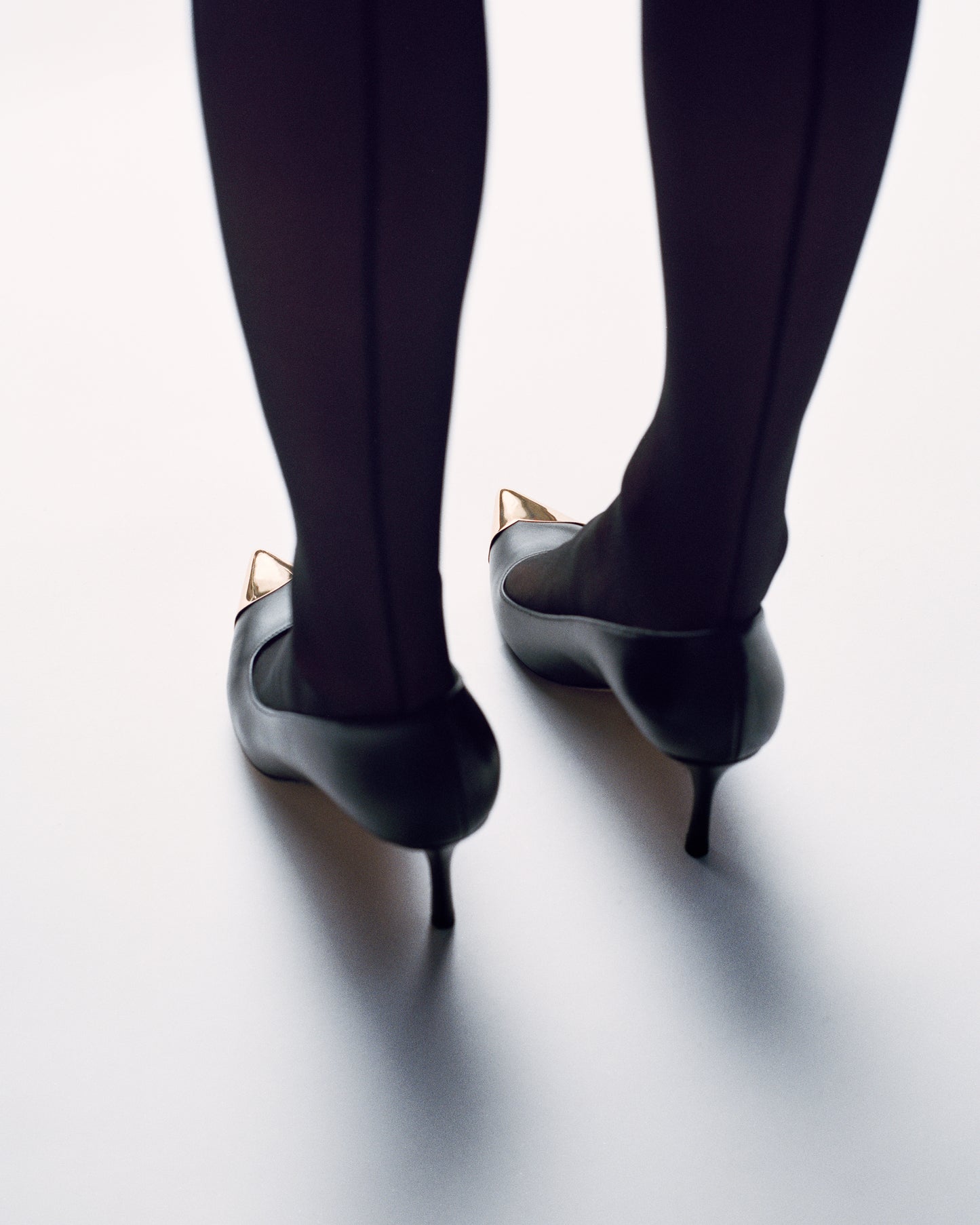 Woman wearing a black leather stiletto heel pump with a gold metallic toe cap and a pointed toe.