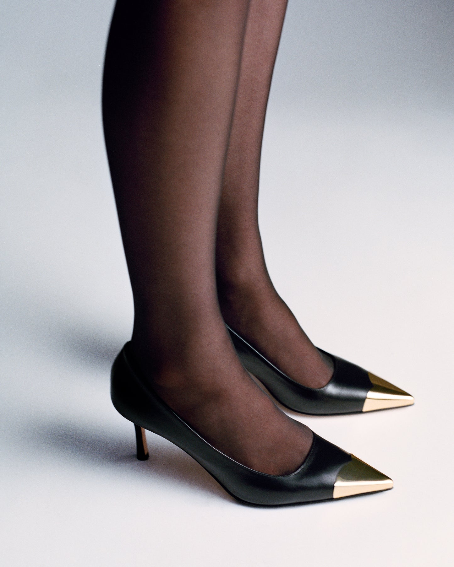 Woman wearing a black leather stiletto heel pump with a gold metallic toe cap and a pointed toe.