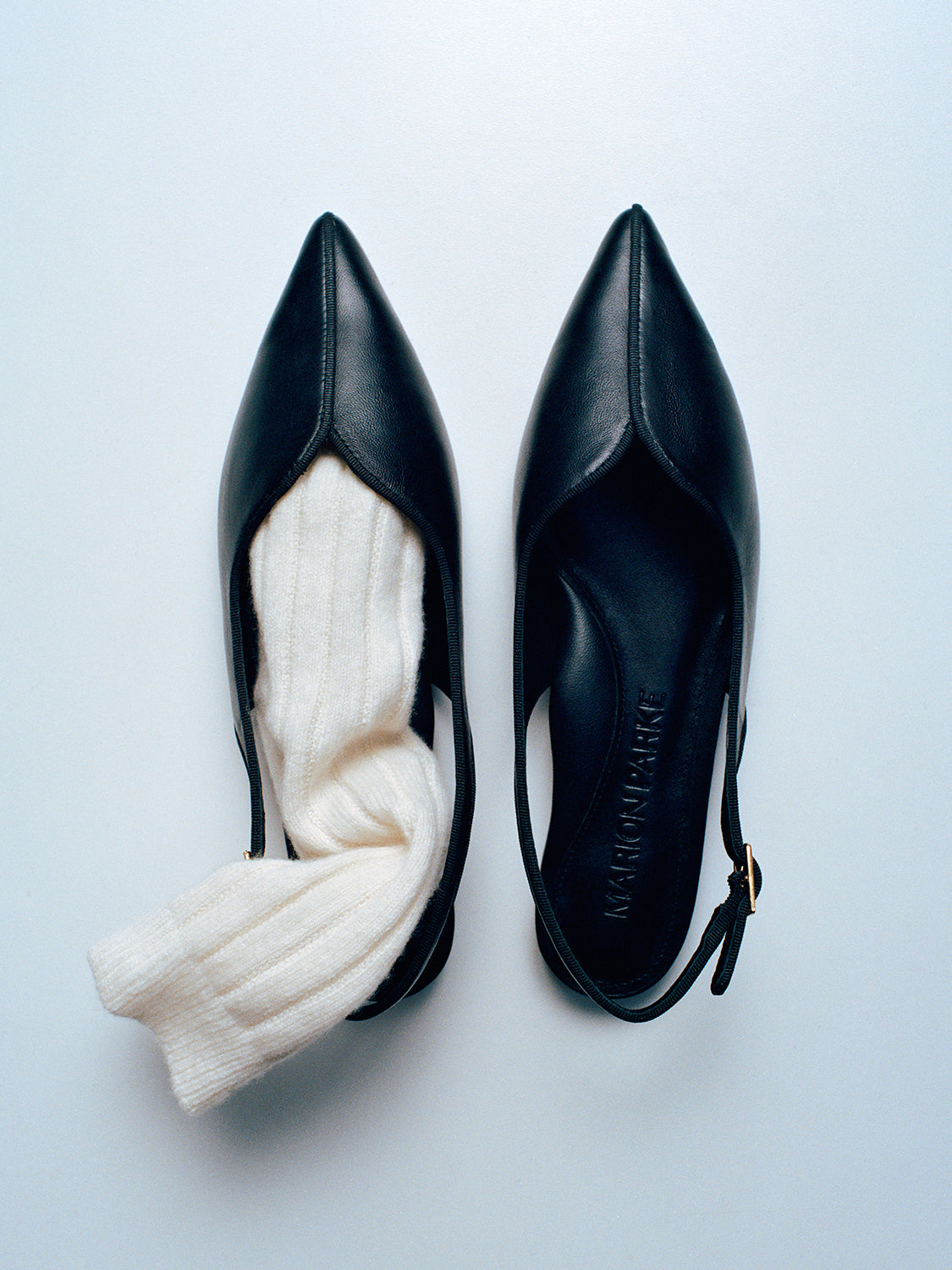 Top view of a pair of black nappa slingback flats with a grosgrain trim detail, styled with a sock.
