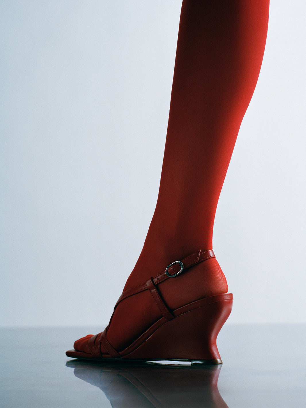 Profile view of a lipstick red leather strappy sandal with a sculptural wedge, shown on a model.