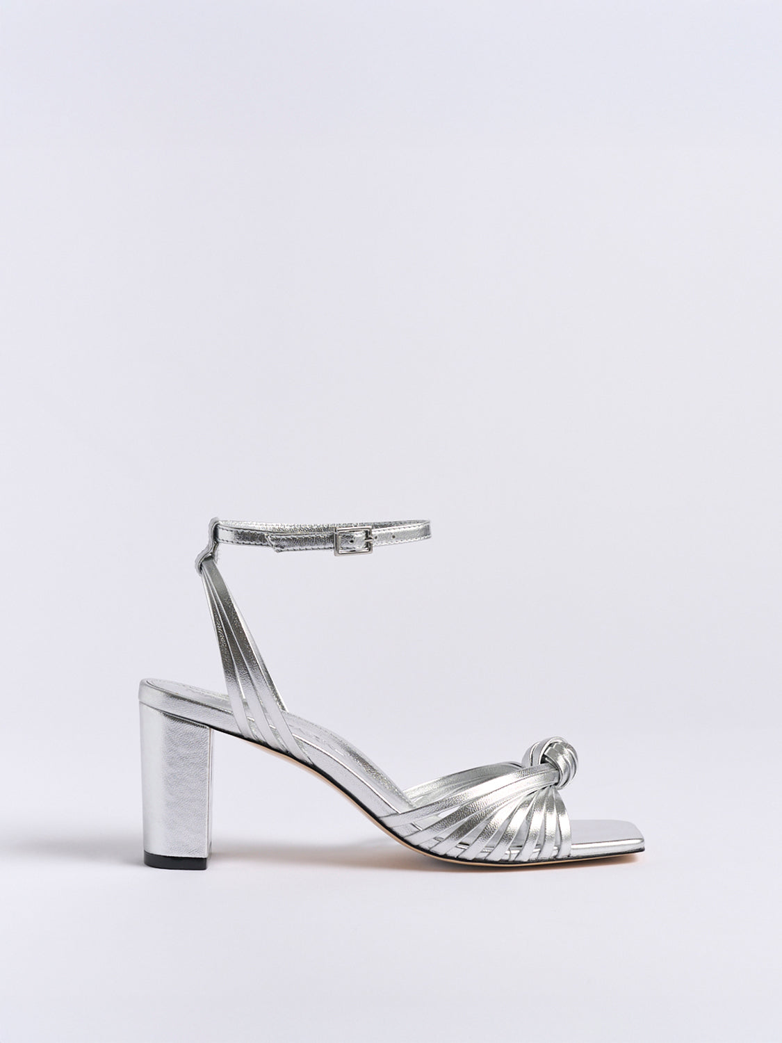 Profile view of a silver metallic leather block heel sandal with a knot detail and a square toe