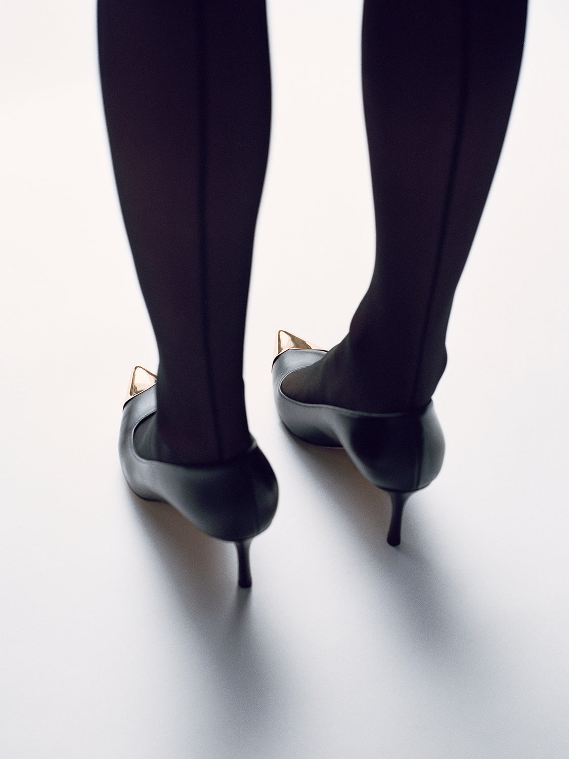 Back view of a black leather pumps with a gold metal toe cap, shown on a model.  