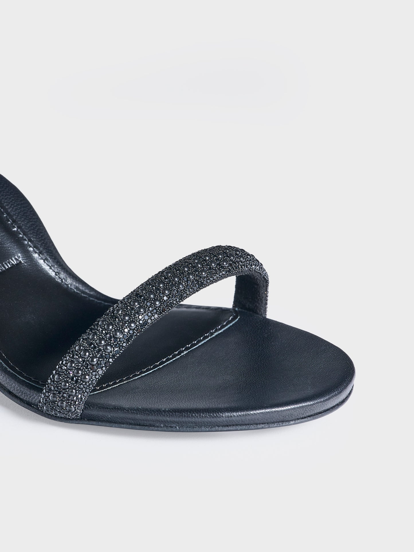 Detail view of a pair of black leather high heel sandals with a crystal embellished toe strap and adjustable ankle strap.