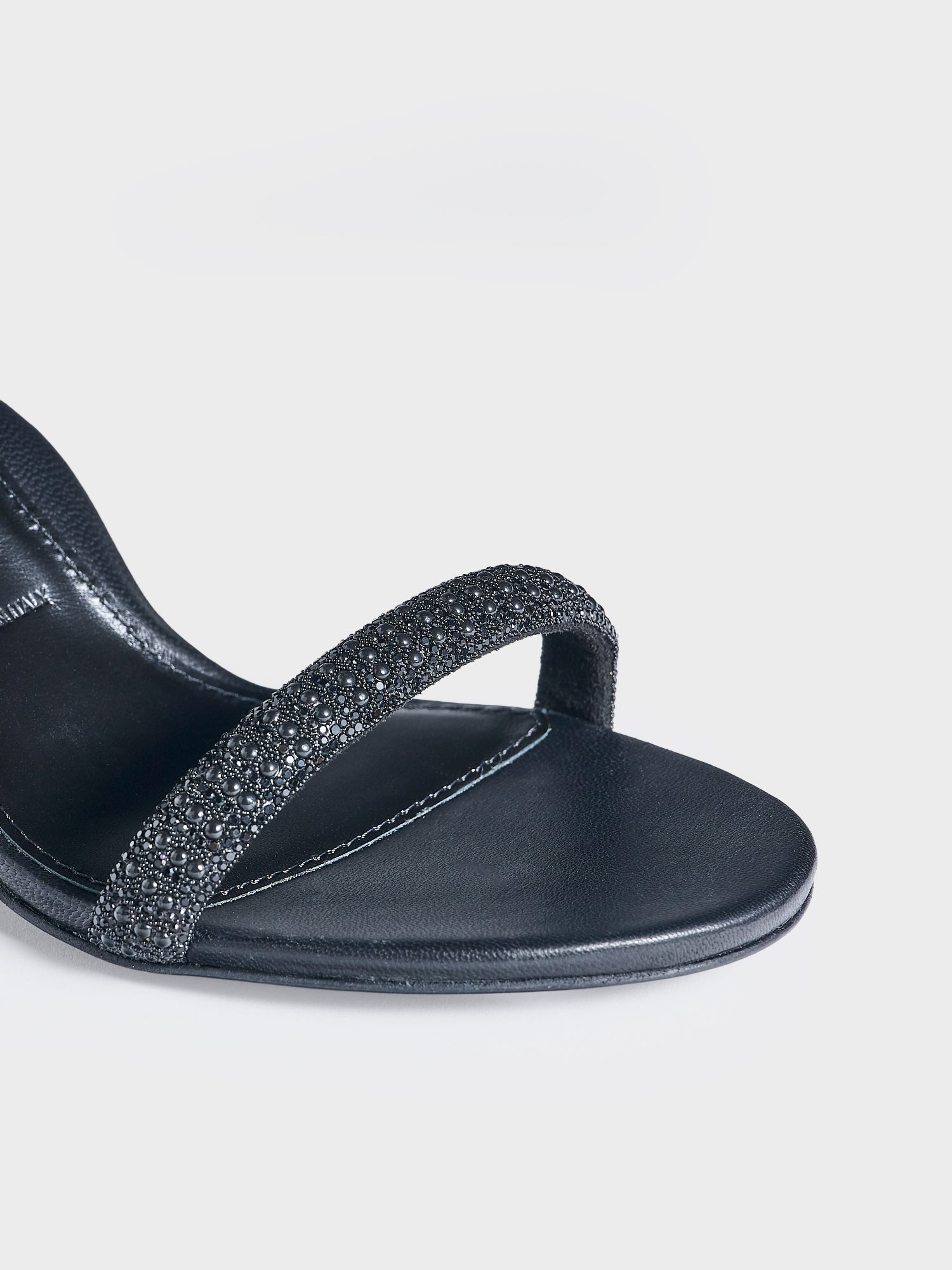 Detail view of a pair of black leather high heel sandals with a crystal embellished toe strap and adjustable ankle strap.