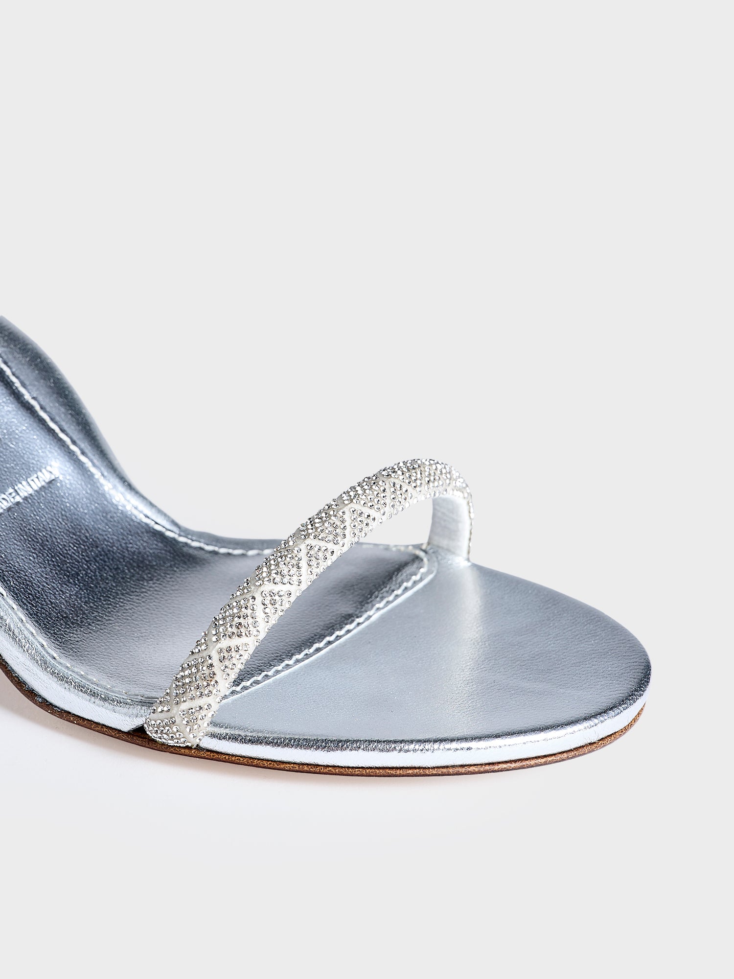Detail view of a pair of silver metallic leather high heel sandals with a crystal embellished toe strap and adjustable ankle strap.