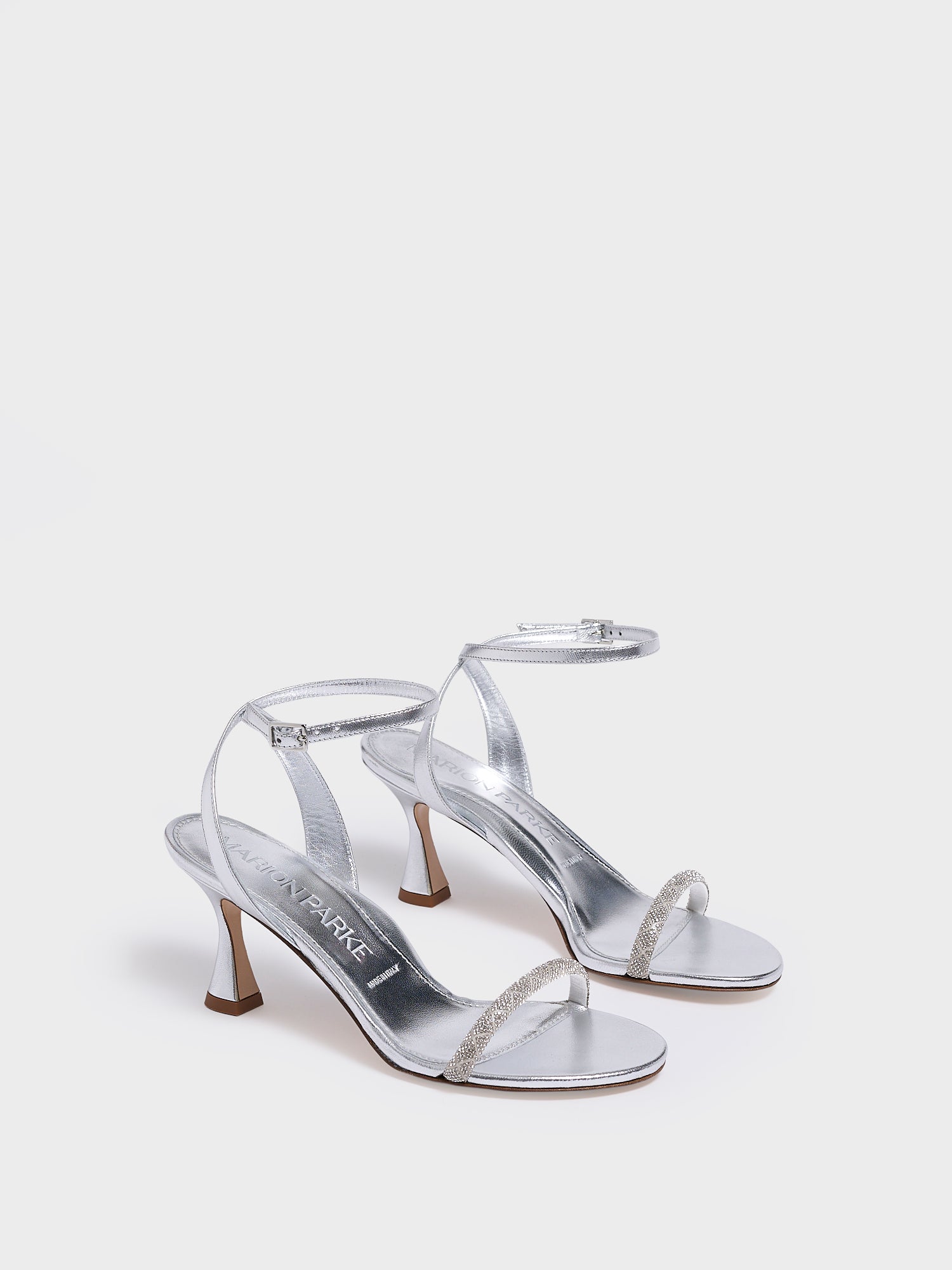 Angle view of a pair of silver metallic leather high heel sandals with a crystal embellished toe strap and adjustable ankle strap.