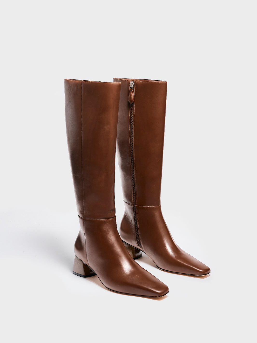 Angle view of a pair of mocha brown nappa leather below the knee tall boots with a rounded heel shape, zipper closure, and square toe. 