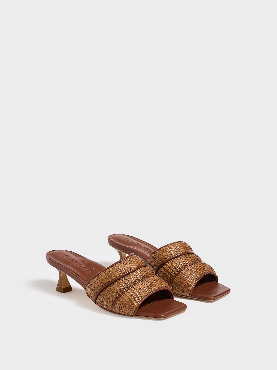 Angle view of a pair of dark brown raffia slide kitten heels with a leather trimmed puffy strap detail.