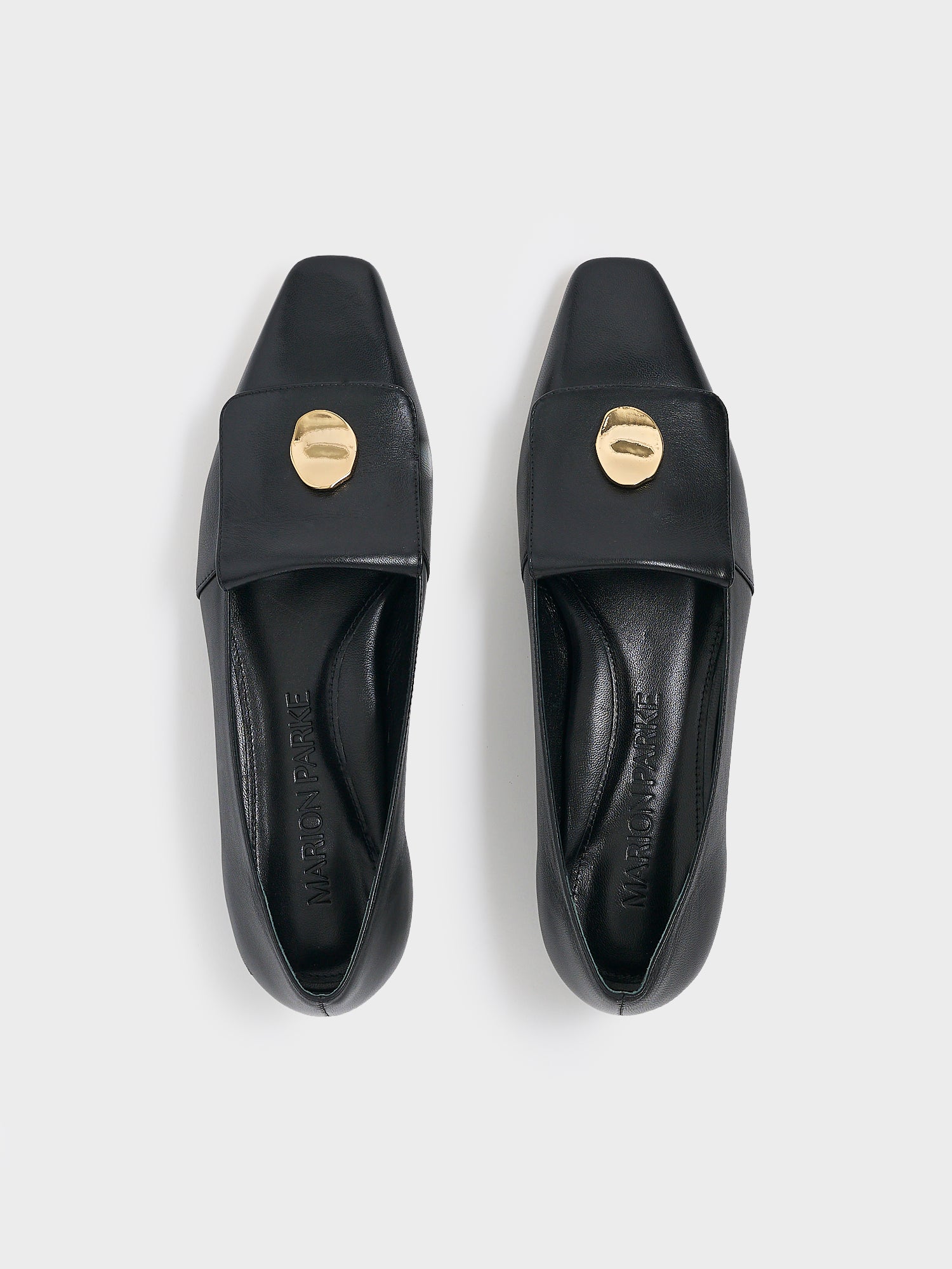 Top view of a pair of black leather flat  loafers with a  gold hardware detail and squared toe. 