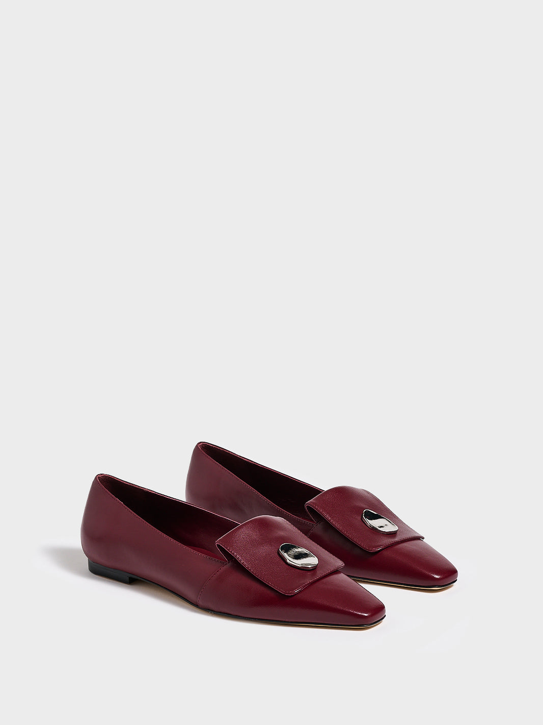 Angle view of a pair of burgundy leather flat  loafers with a  silver hardware detail and squared toe. 