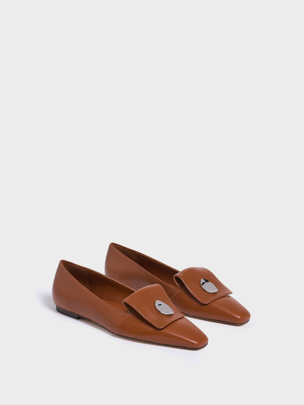 Angle view of a pair of chestnut leather  flat loafers with a  silver hardware detail and squared toe. 