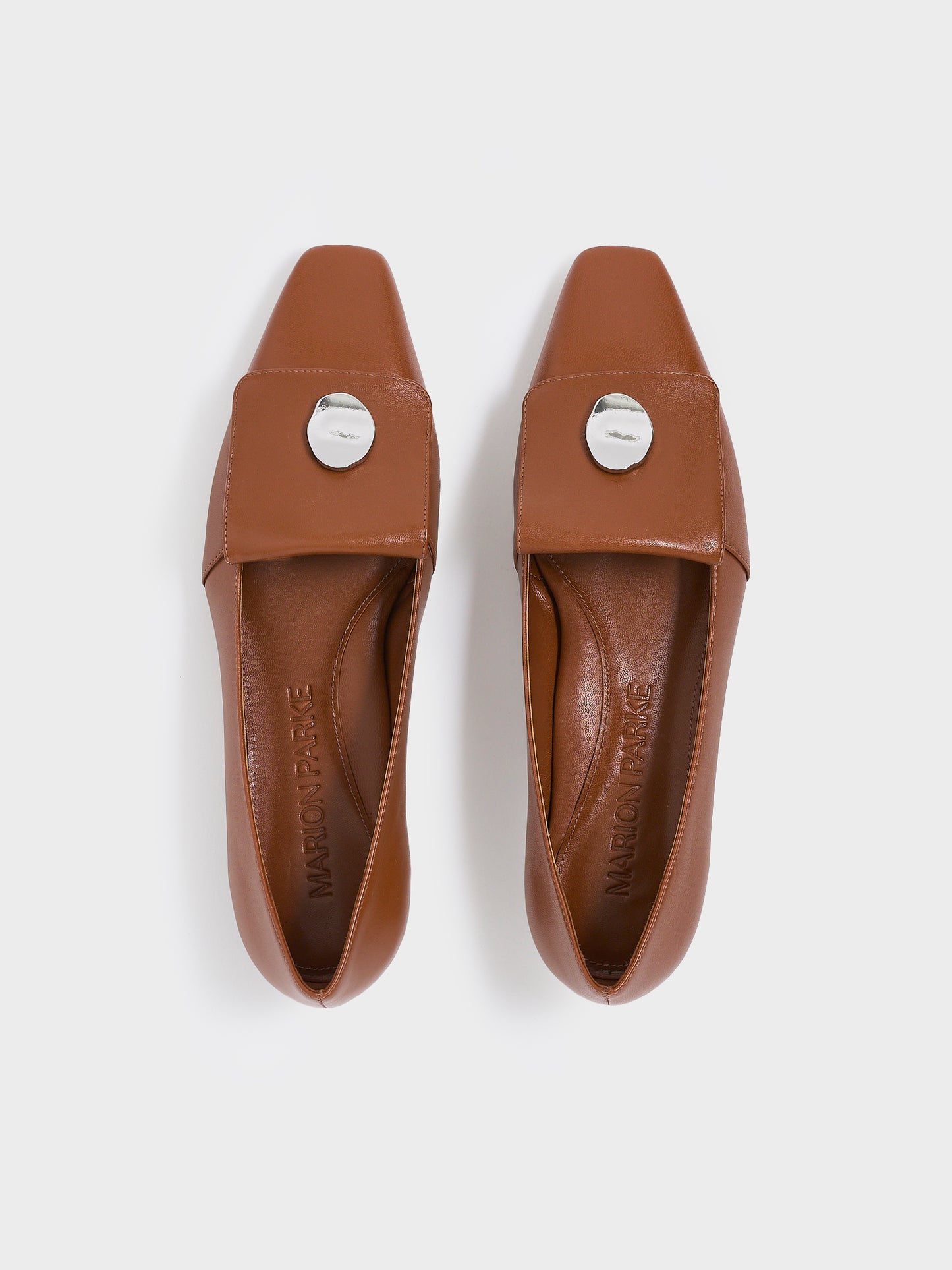 Top view of a pair of chestnut leather  flat loafers with a silver hardware detail and squared toe. 
