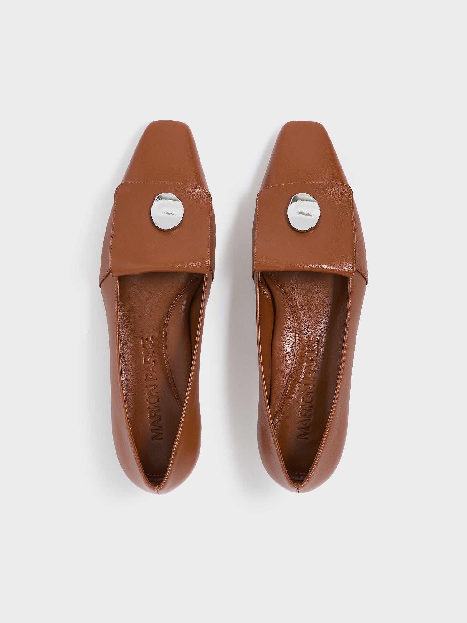 Top view of a pair of chestnut leather  flat loafers with a silver hardware detail and squared toe. 