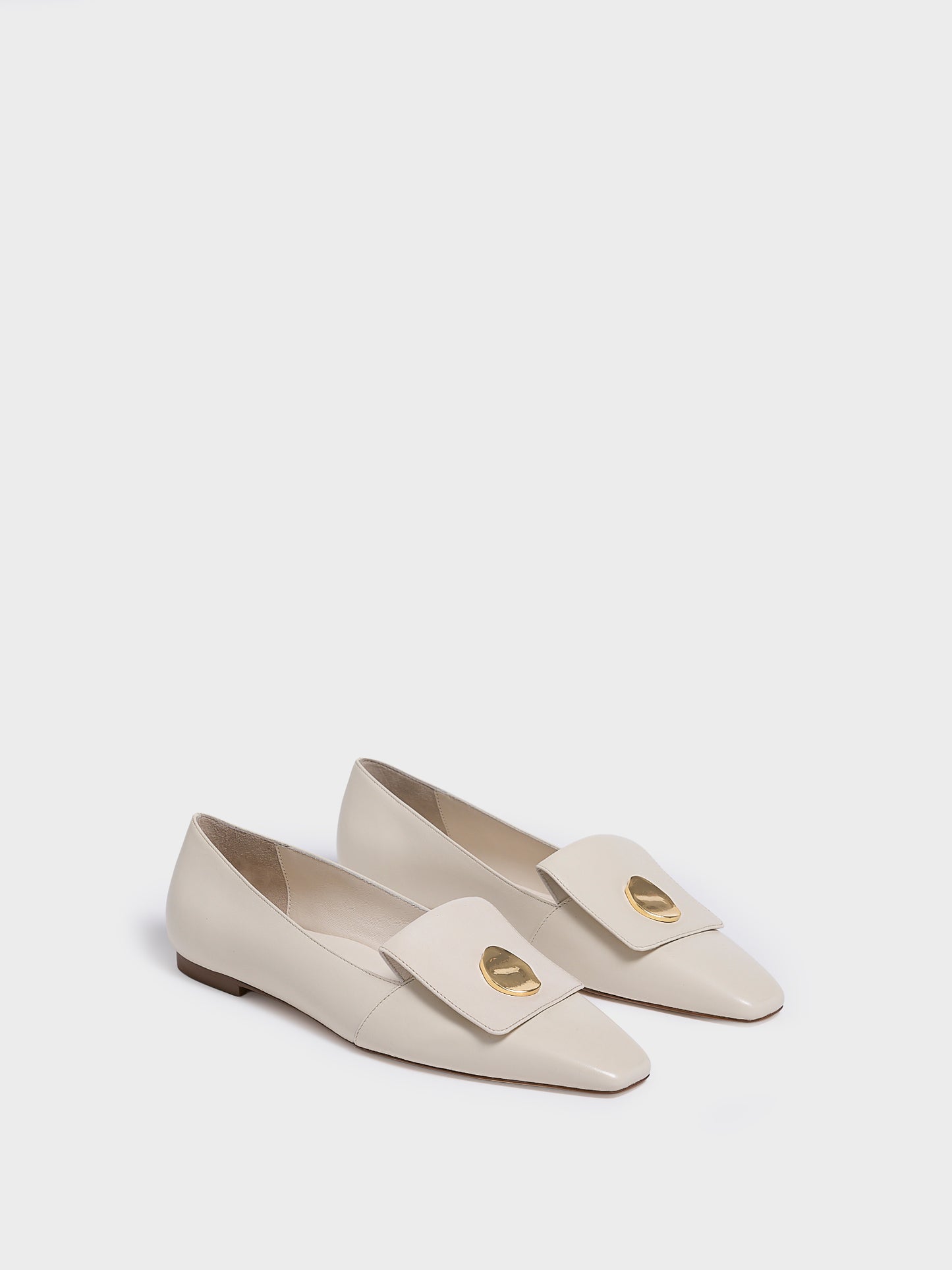 Angle view of a pair of ivory leather  flat loafers with a  gold hardware detail and squared toe. 